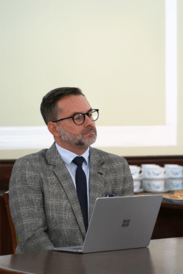 Mężczyzna w szarym garniturze i okularach prowadzi prezentację, siedząc przy laptopie podczas spotkania w sali konferencyjnej.