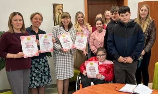 Grupa osób pozuje do zdjęcia w pomieszczeniu, trzymając plakaty promujące kiermasz ciast na rzecz Fundacji Cancer Fighters.